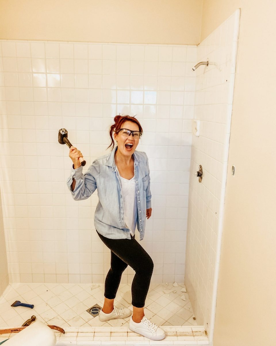 Anita, hammer in hand, about to demo a white tile bathroom