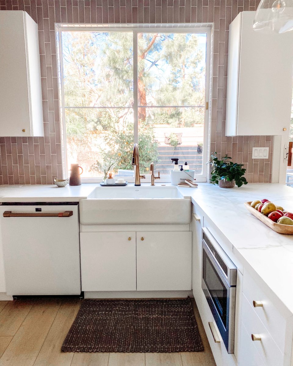 A white farmhouse sink below a bright window set in white marble with a brass Delta faucet