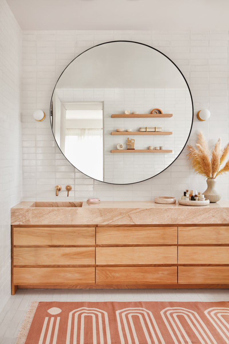 Natural earthen counter set atop wooden vanity with large round mirror backed by white fireclay tile