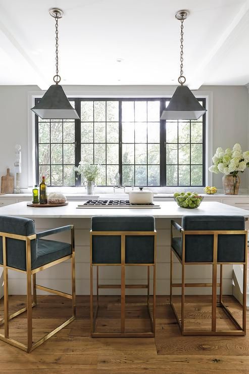 Blue velvet counter stools sit at a white shiplap island topped with a honed white marble countertop holding a gas integrated cooktop beneath black metal vintage lights.