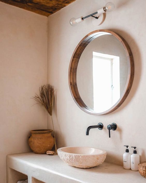 Earthen vessel sink set below a round wooden mirror with black matte faucet and fixtures