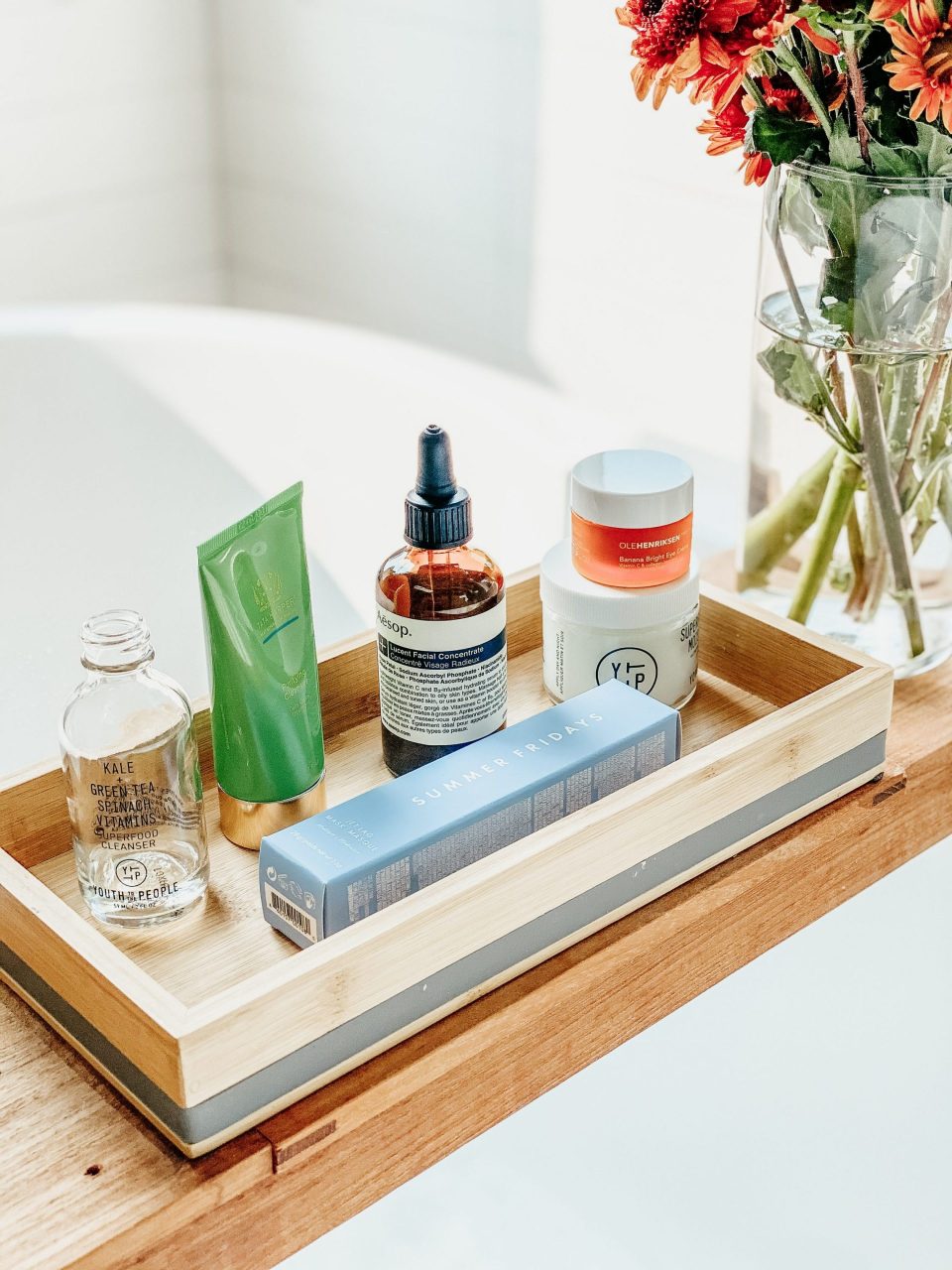 Array of beauty products on a bath tray next to a vase of orange flowers
