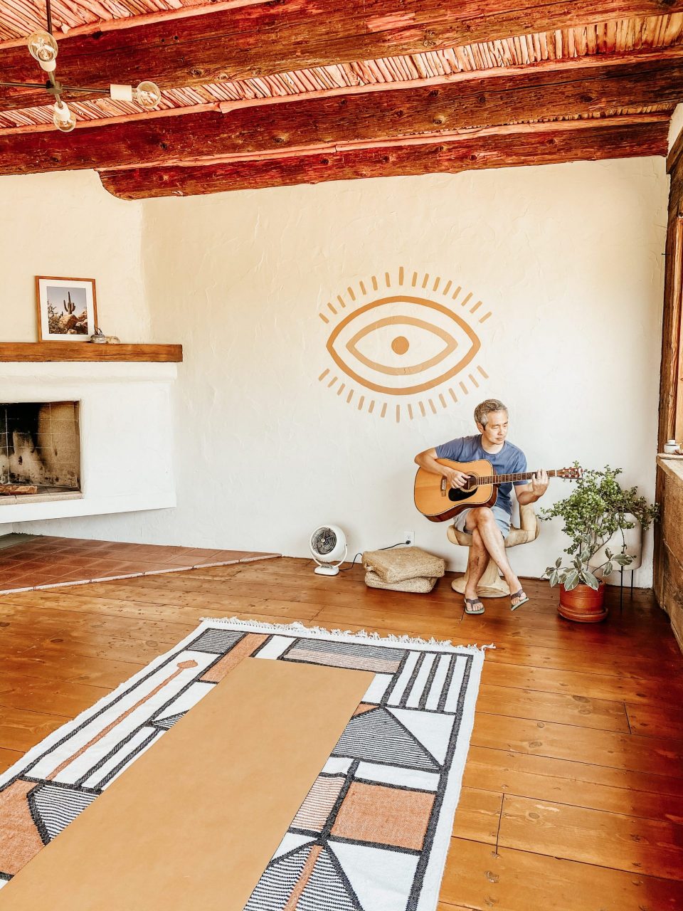 Travis playing guitar in an open yoga room beneath a minimalist mural of an eye